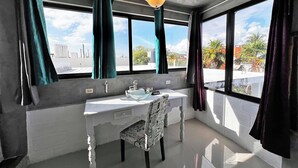 Ein Badezimmer mit einem weißen Waschbecken, einem Spiegel und einem Stuhl. Zwei Fenster mit Vorhängen und eine Deckenleuchte sind vorhanden.