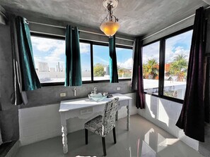 Ein Badezimmer mit einem weißen Waschbecken, einem Spiegel und einem Stuhl. Zwei Fenster mit Vorhängen und eine Deckenleuchte sind vorhanden.