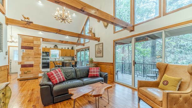 A spacious living area with a black leather sofa, a wooden coffee table, and a tan armchair. A kitchen is visible in the background with wooden cabinets and a large window. - Heart of Helen Cabin (Helen)