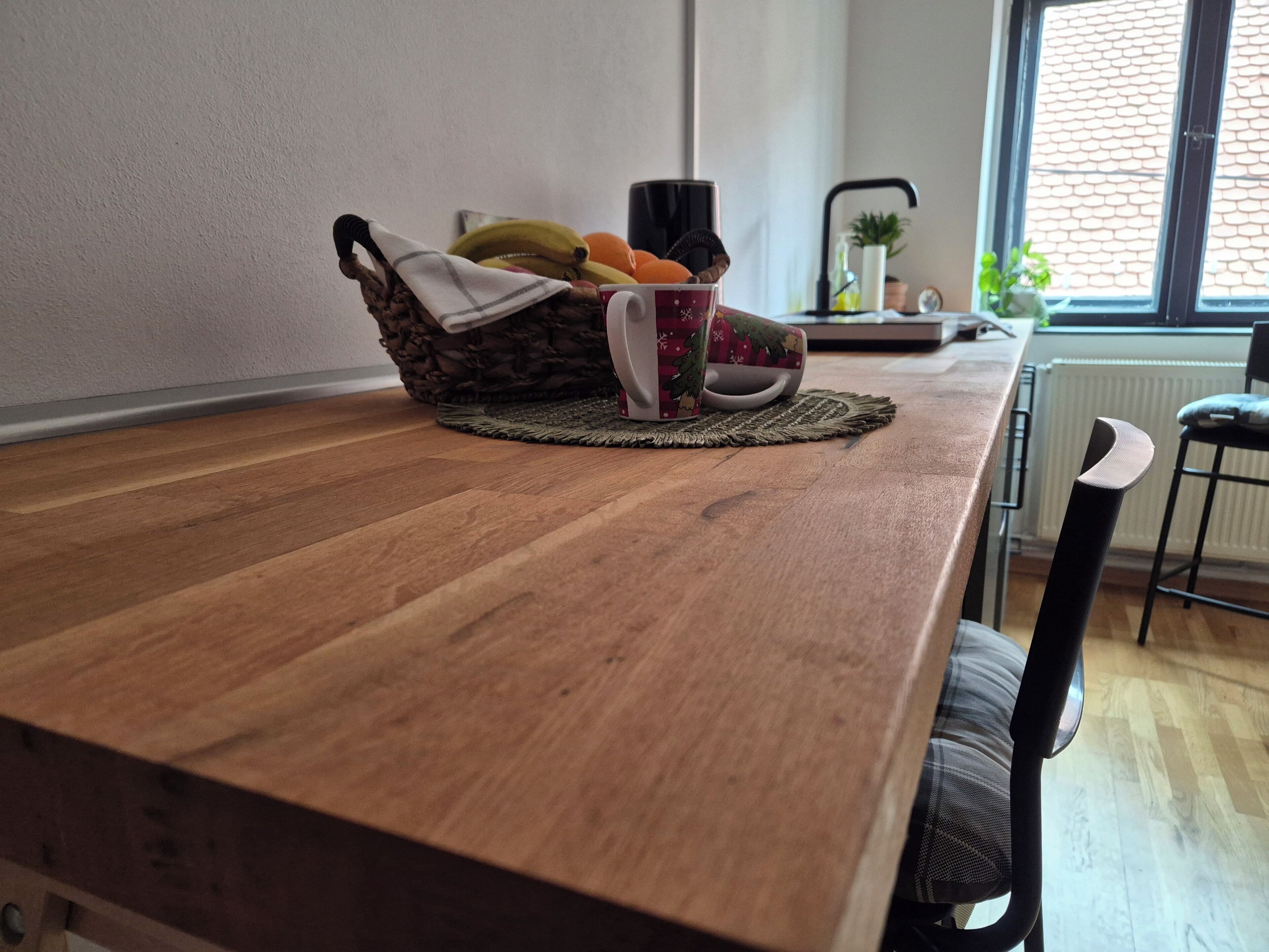 Une table de salle à manger en bois, surmontée d’un set de table en fibres tressées, un panier rempli de fruits et une tasse, une chaise, et une fenêtre donnant sur un mur de briques.