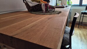 Une table de salle à manger en bois, surmontée d’un set de table en fibres tressées, un panier rempli de fruits et une tasse, une chaise, et une fenêtre donnant sur un mur de briques.