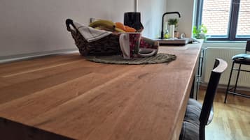 Une table de salle à manger en bois, surmontée d’un set de table en fibres tressées, un panier rempli de fruits et une tasse, une chaise, et une fenêtre donnant sur un mur de briques.