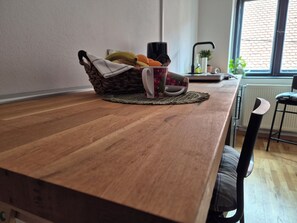 Ein Holztisch mit einer gewebten Tischmatte, ein Korb mit Früchten und einer Tasse, ein Stuhl und ein Fenster mit Blick auf eine Ziegelwand.