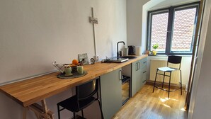 Une cuisine avec un plan de travail en bois, des chaises noires, un luminaire suspendu, une fenêtre donnant sur un toit recouvert de tuiles rouges et un radiateur.