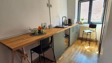 Une cuisine avec un plan de travail en bois, des chaises noires, un luminaire suspendu, une fenêtre donnant sur un toit recouvert de tuiles rouges et un radiateur.