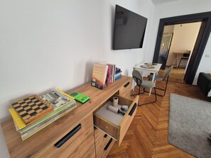Ein Zimmer mit einem Holztisch, Büchern, einem Flachbildfernseher, einem kleinen Tisch mit Stühlen und einem Fischgrätparkett.