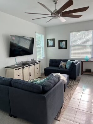 Ein Wohnzimmer mit Deckenventilator, Flachbildfernseher, einem dunkelgrauen Sofa, einem Couchtisch und einer Kommode mit Schubladen.
