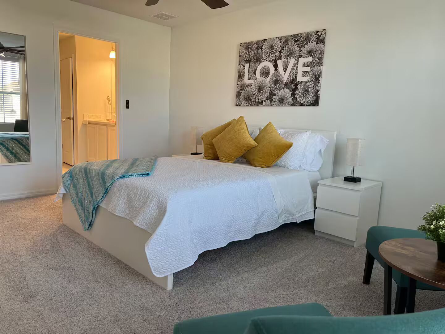 A bedroom with a bed, a nightstand, a bedside table, a chair, a small table with a plant, and a wall with a 'LOVE' sign.