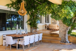 An outdoor dining area with a wooden table and chairs, a hanging light fixture, and a tree providing shade.