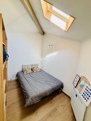 Ein Schlafzimmer mit schrÀgen WÀnden, einem Bett mit floralen Kissen, einem Holzschrank und einem Schminktisch mit Spiegel und Kartenablage.