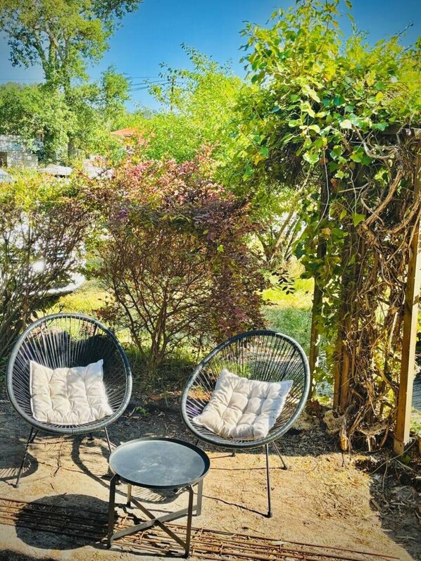 Zwei KorbstĂŒhle mit Kissen, ein kleiner runder Tisch und ein Garten mit BĂ€umen und einer Holzkonstruktion.