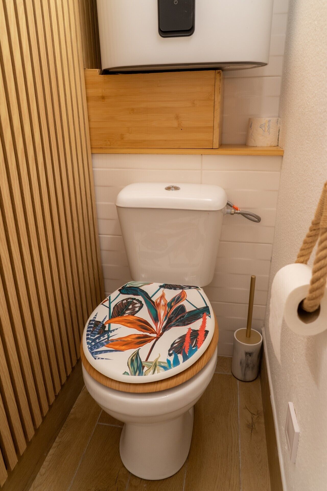 Un inodoro con un asiento de motivos florales, un panel de madera encima y un rollo de papel higiénico en un estante.