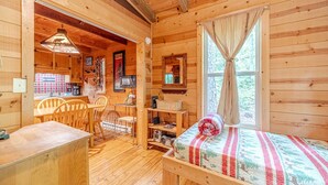 A wooden cabin interior with a dining area, a bed with a patterned blanket, and a window with curtains. - Unicoi Pines | Hot Tub, Pet-Friendly Cabin Near Helen, GA (Sautee Nacoochee)