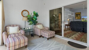 Ein Wohnzimmer mit gestreiften Sesseln, einem Beistelltisch mit Lampe, einer großen Grünpflanze und einer Uhr an der Wand.