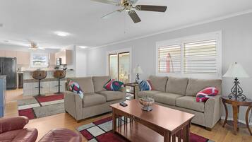 A living room with a sofa, a coffee table, a red leather chair, a ceiling fan, and a kitchen in the background.