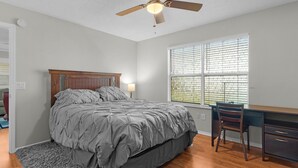 Um quarto com cama, escrivaninha, cadeira, ventoinha no teto e janela com persianas.