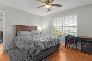 Ein Schlafzimmer mit einem Bett, einem Schreibtisch, einem Stuhl, einem Deckenventilator und einem Fenster mit Jalousien.