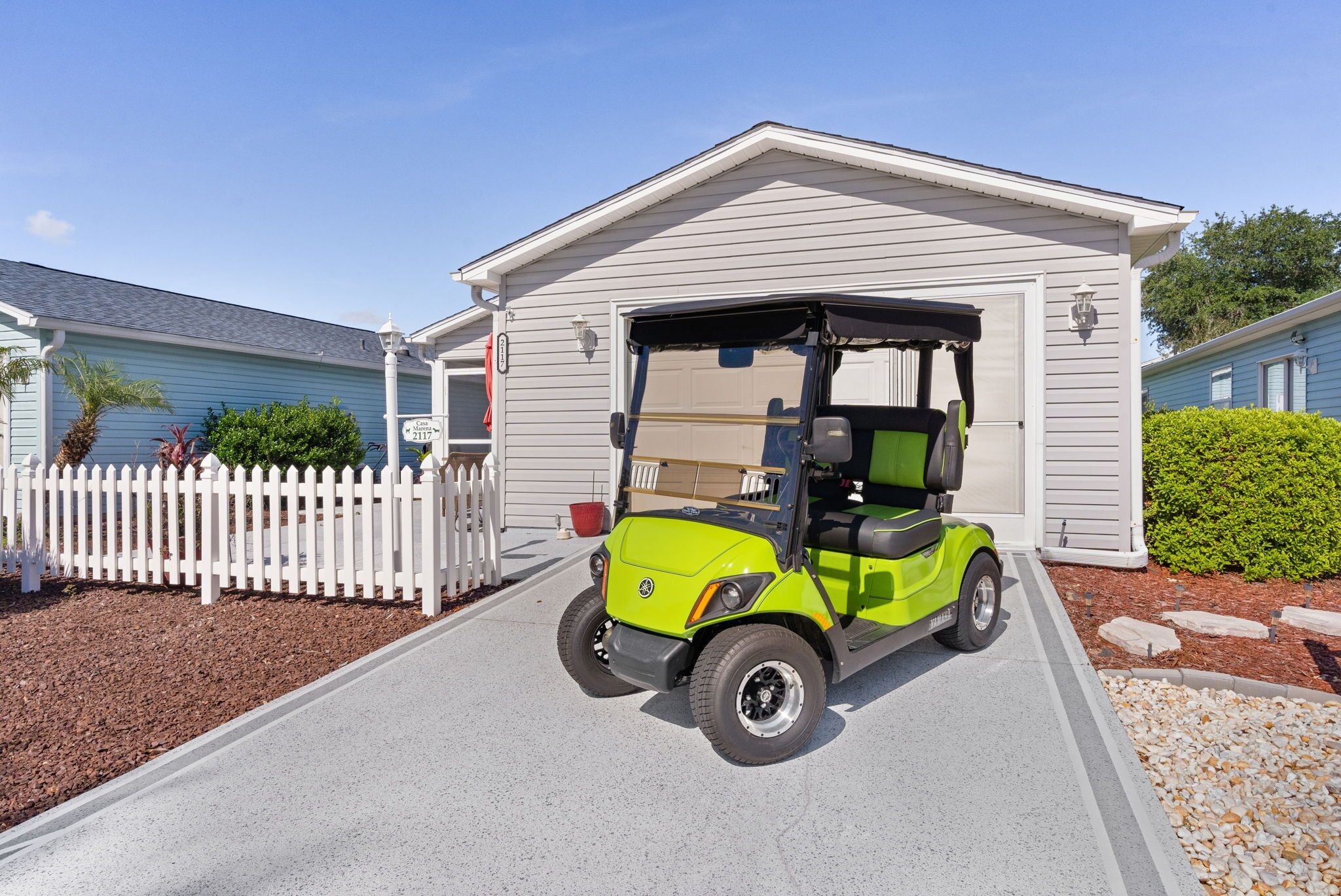 A golf cart parked in front of a building with a garage door, a white picket fence, and a clear sky.