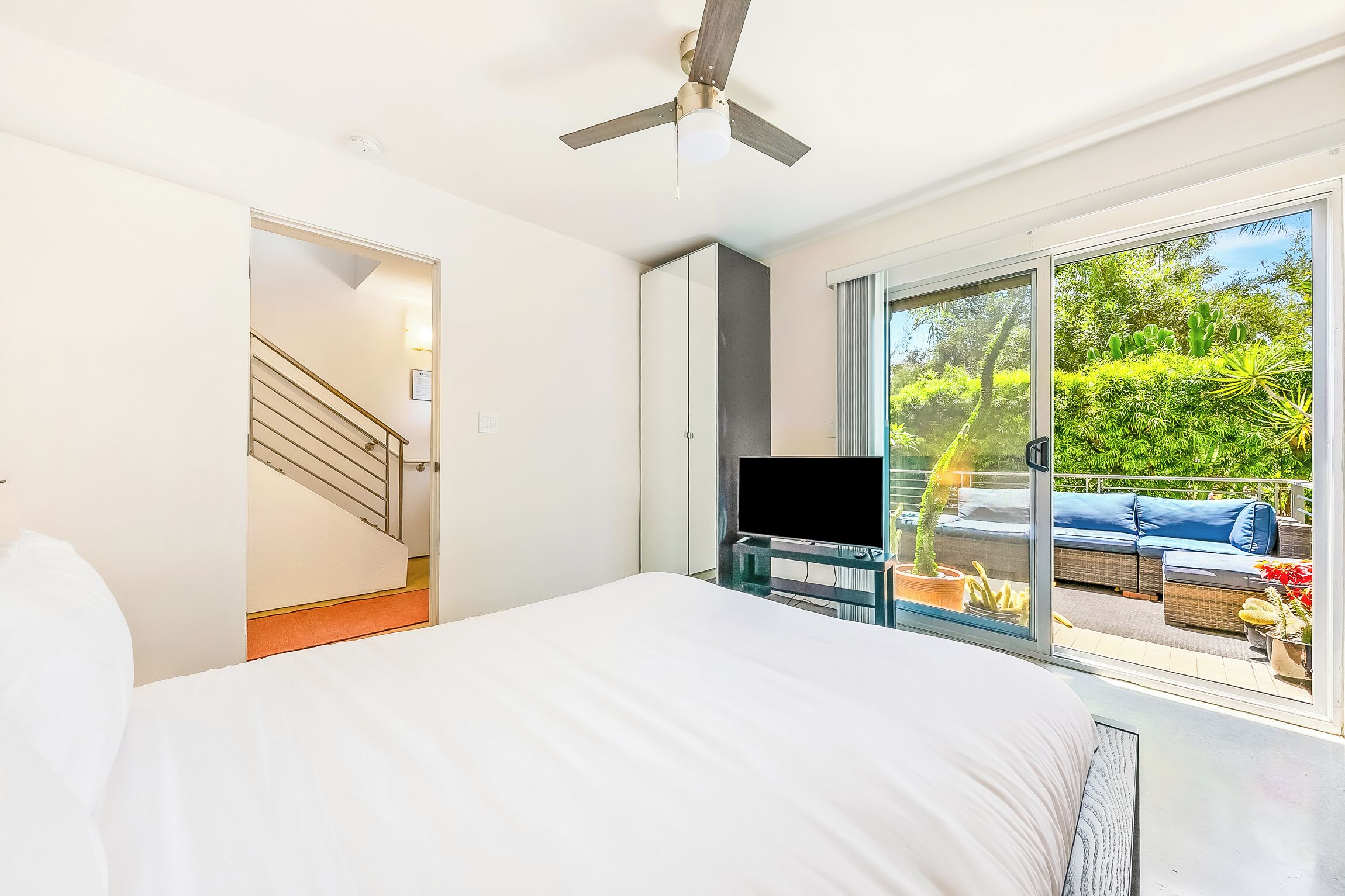 A bedroom with a bed, a TV on a stand, a ceiling fan, and a sliding glass door leading to an outdoor seating area with a couch and plants.