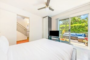 Une chambre avec un lit, un téléviseur posé sur un meuble, un ventilateur de plafond et une porte-fenêtre coulissante donnant sur un espace extérieur aménagé avec un canapé et des plantes.