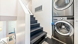 Uma lavanderia com máquina de lavar e secadora empilhadas, escada e um duto na parede.