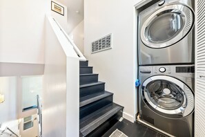 A laundry room with a stacked washer and dryer, a staircase, and a vent on the wall. - Heart Of North Park, Mins To Beach & Restaurants (San Diego)