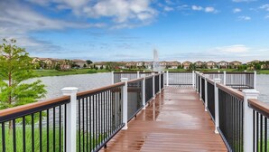 A wooden deck leading to a body of water with a fountain, surrounded by houses. - Fun Family Townhome with Private Pool & Game Room! (Kissimmee)