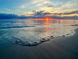 Eine Strandansicht mit Fußspuren, die zum Wasser führen, einem Sonnenuntergang und einer ruhigen See.