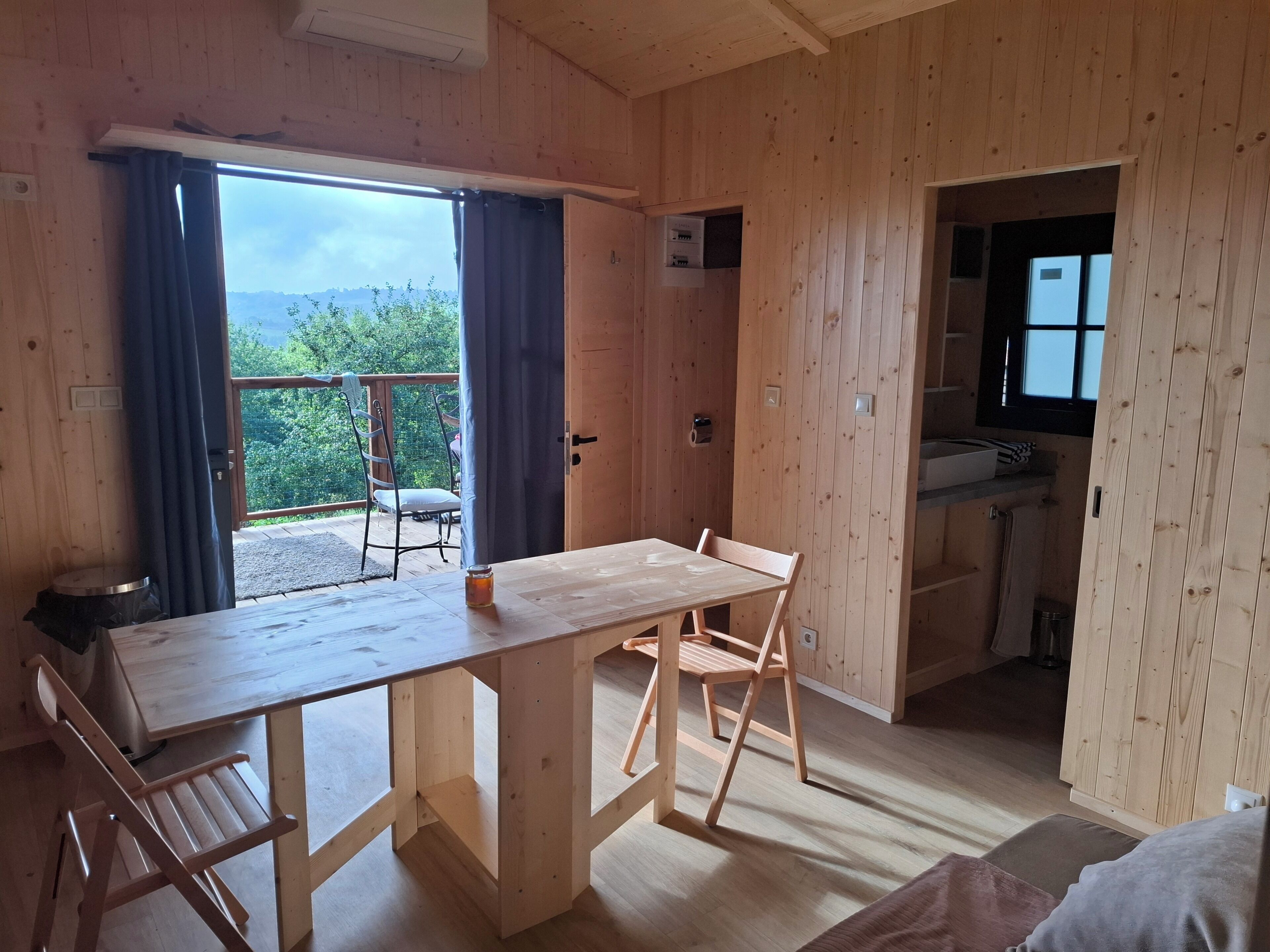 Un intérieur en bois comprenant une table à manger, des chaises, un petit coin cuisine et une vue sur l’extérieur, visible à travers une porte.