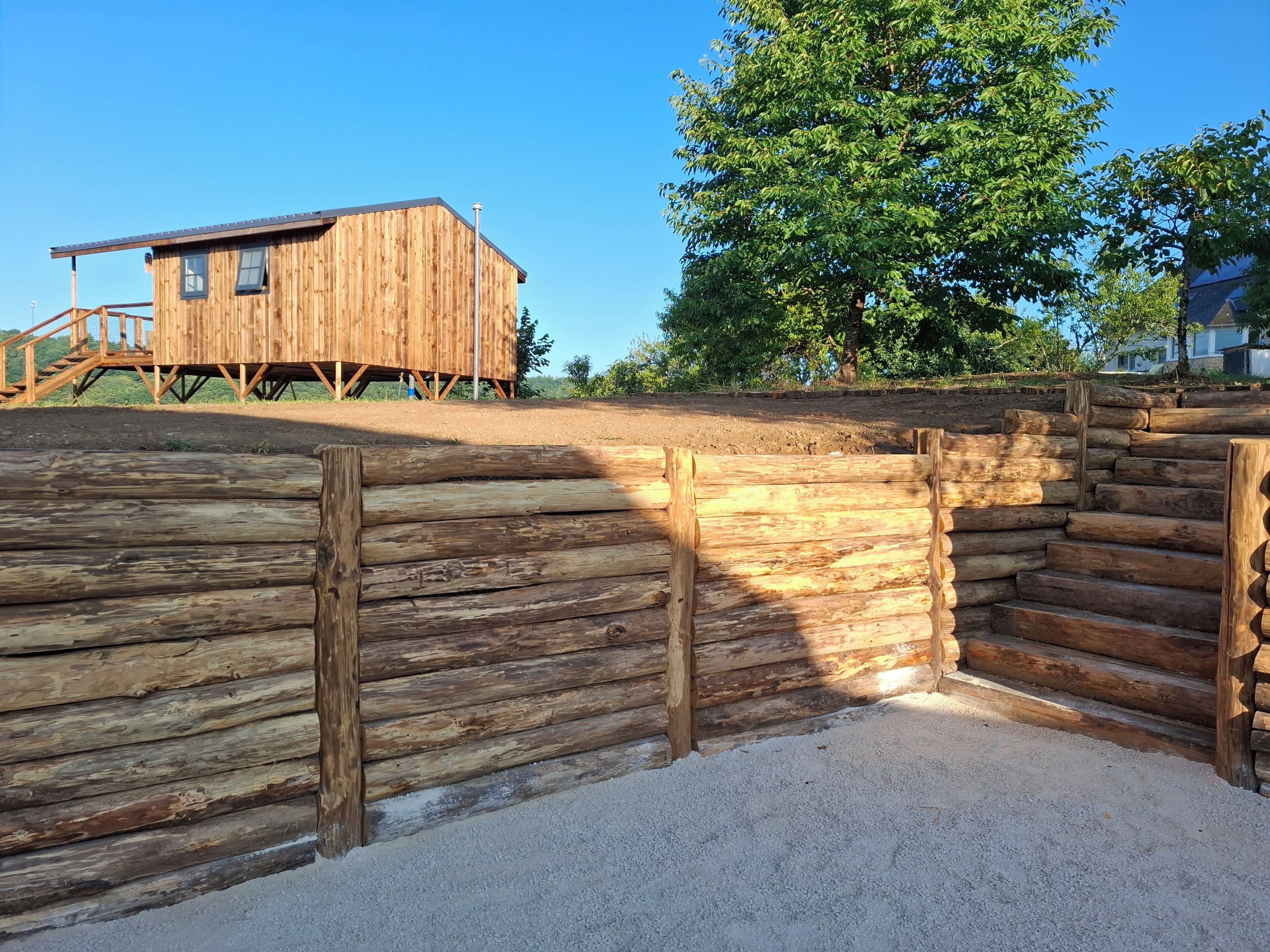 Een houten blokhut met een trap die erheen leidt, omringd door een houten hek en een boom.