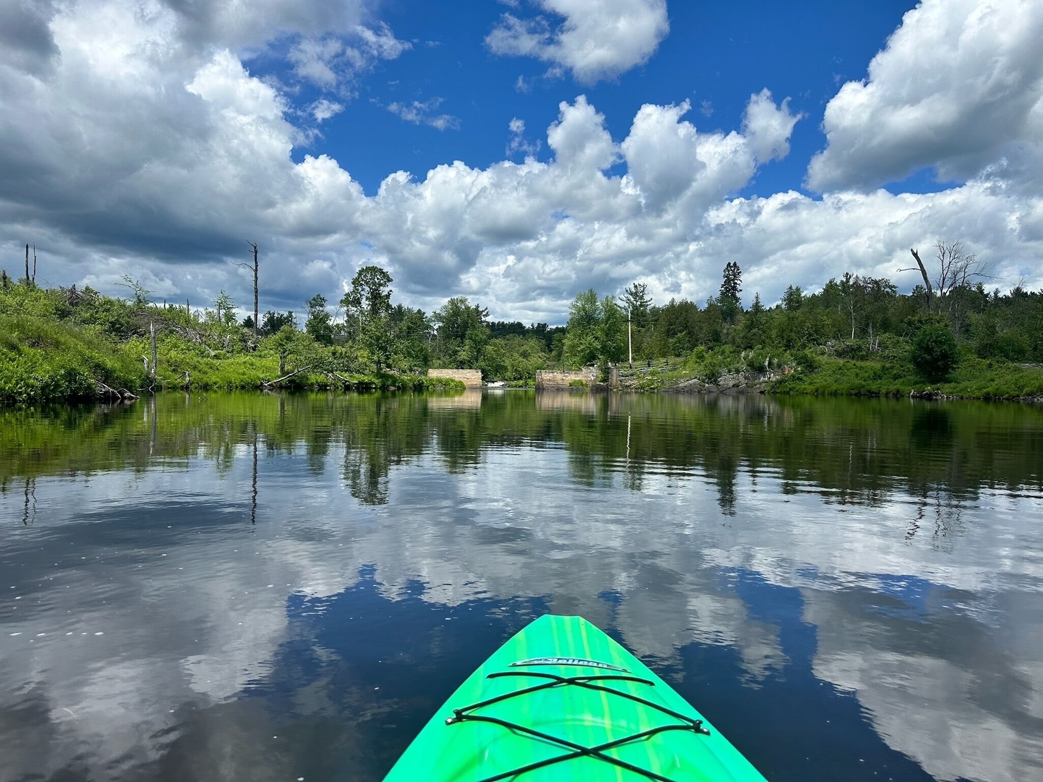 Un río tranquilo con un kayak verde en primer plano, rodeado de exuberante vegetación y árboles bajo un cielo parcialmente nublado.