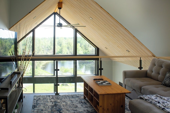 Un dormitorio estilo loft moderno con techo inclinado, ventanas amplias, un escritorio de madera, un sofá y un televisor.