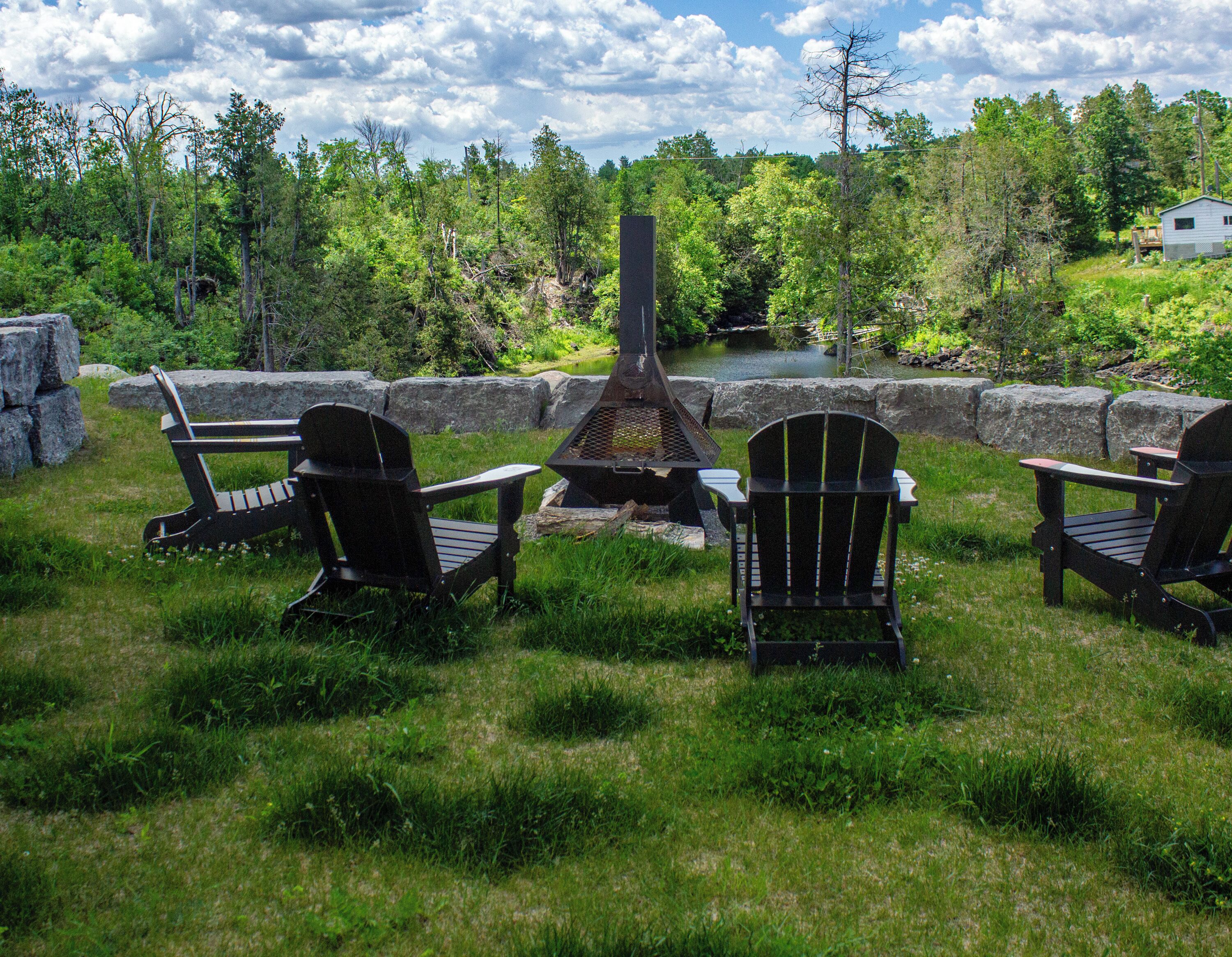Un espace extérieur verdoyant avec un foyer en pierre, trois fauteuils Adirondack et un étang en arrière-plan.