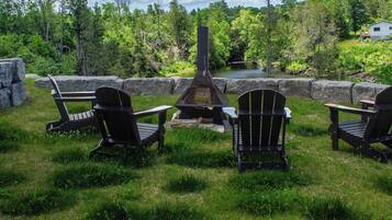 Un espace extérieur verdoyant avec un foyer en pierre, trois fauteuils Adirondack et un étang en arrière-plan.