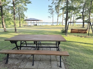 Une table de pique-nique avec des bancs, située dans un parc arboré, avec un kiosque en arrière-plan.