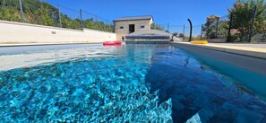 Une piscine aux eaux cristallines bordée de pierres, un petit bâtiment en arrière-plan et une clôture qui délimite l’espace.