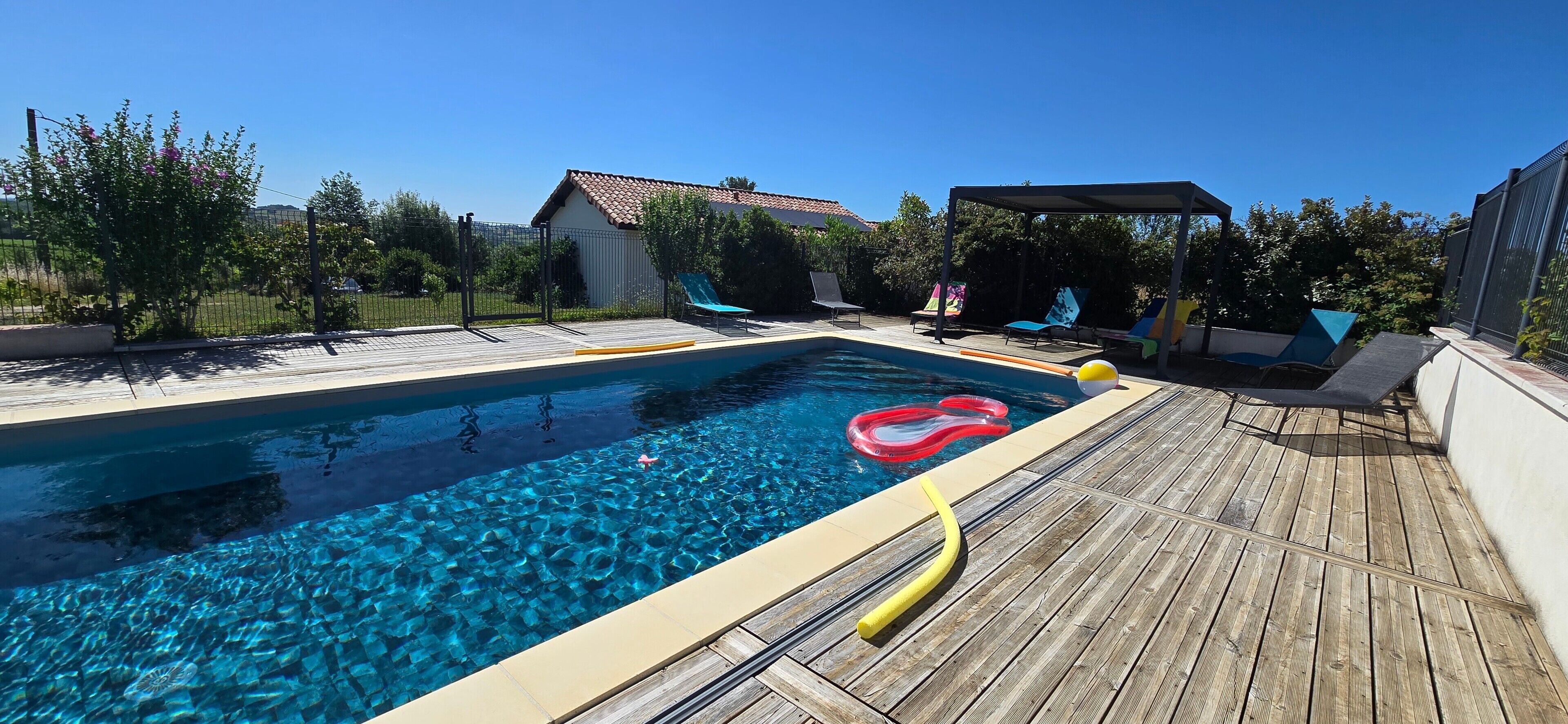 Piscina com deck de madeira, área de estar coberta e jardim cercado.