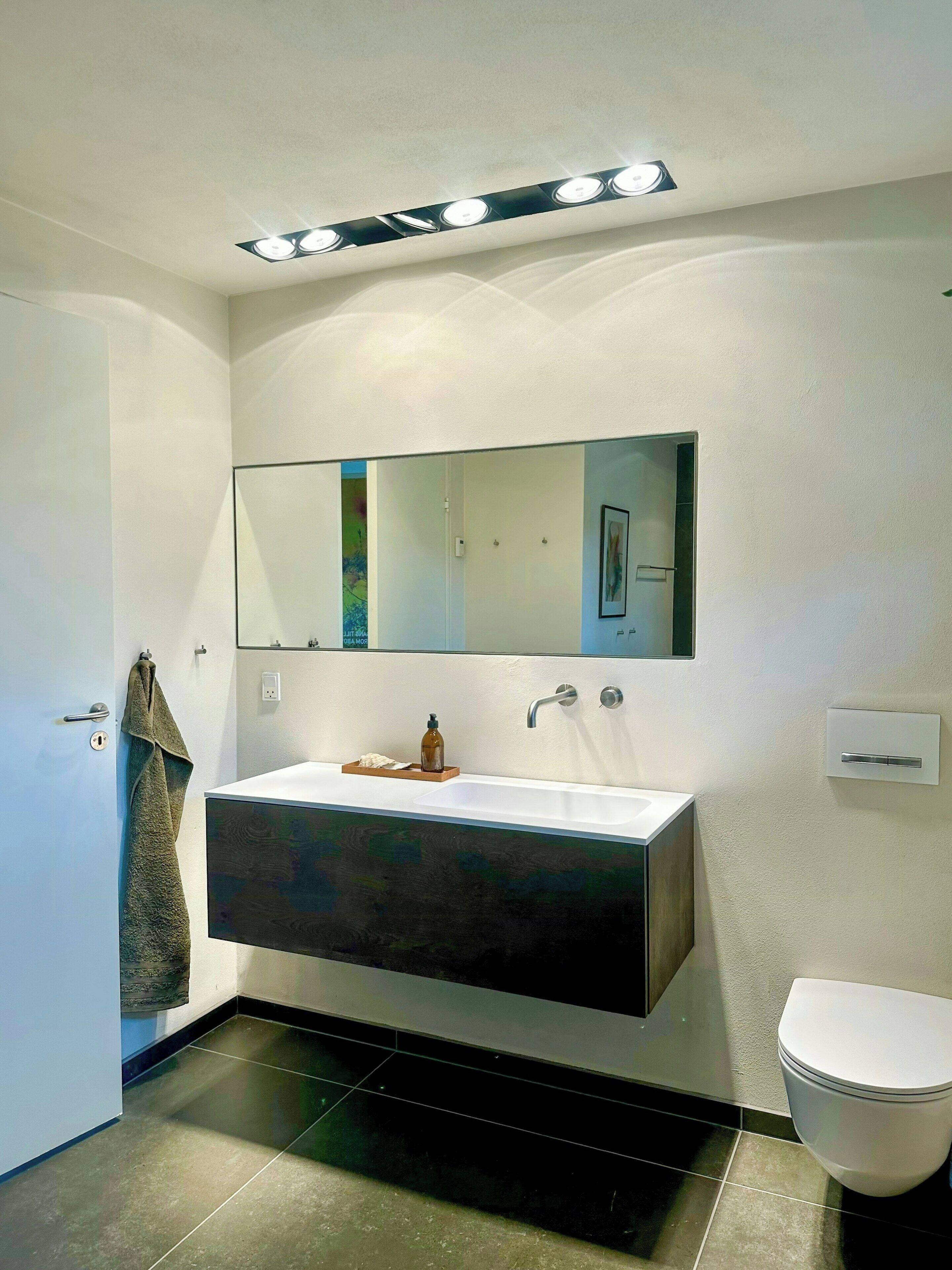 Uma casa de banho moderna com um espelho grande, uma sanita, uma pia branca, um lavatório preto e uma toalha pendurada na porta.