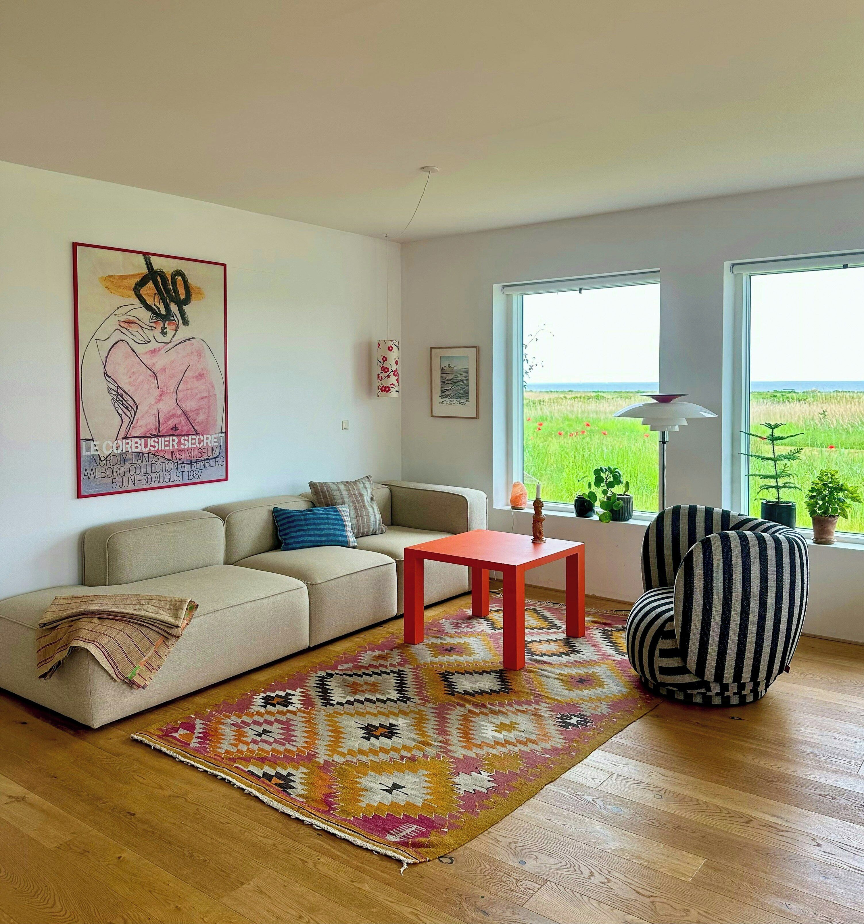 Un salon avec un canapé, une table rouge, un tapis à motifs, une chaise à rayures et une grande fenêtre donnant sur un champ.