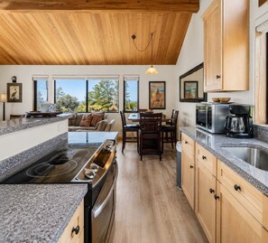 A modern kitchen with granite countertops, wooden cabinets, and a dining area with a view. - Love Nest | Ocean View Cottage & Hot Tub (Sea Ranch)