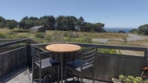A balcony with a round table and chairs, overlooking a coastal landscape with trees and a clear sky. - Love Nest | Ocean View Cottage & Hot Tub (Sea Ranch)