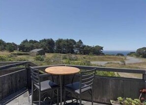 A balcony with a round table and chairs, overlooking a coastal landscape with trees and a clear sky. - Love Nest | Ocean View Cottage & Hot Tub (Sea Ranch)