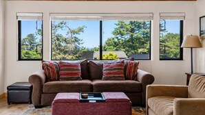 A living room with a sofa, ottoman, and armchair, large windows with a view of trees, and a lamp on the right. - Love Nest | Ocean View Cottage & Hot Tub (Sea Ranch)