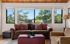A living room with a sofa, ottoman, and armchair, large windows with a view of trees, and a lamp on the right. - Love Nest | Ocean View Cottage & Hot Tub (Sea Ranch)