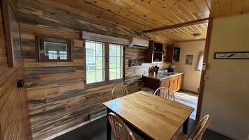 A rustic cabin interior with a wooden dining table, chairs, a kitchen area with cabinets, a window, and a door.