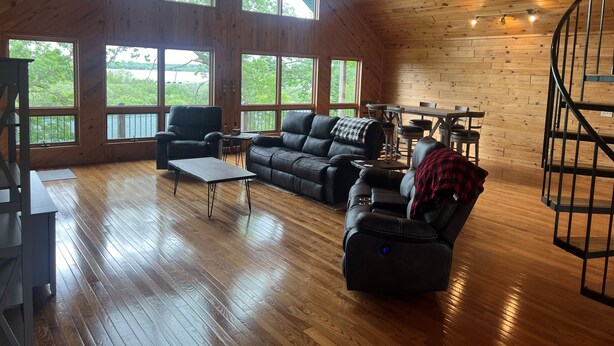 A spacious living room with wooden floors, large windows, a black leather sofa, a recliner, and a dining area with a table and chairs. - Cass Lake Lodge (Cass Lake)
