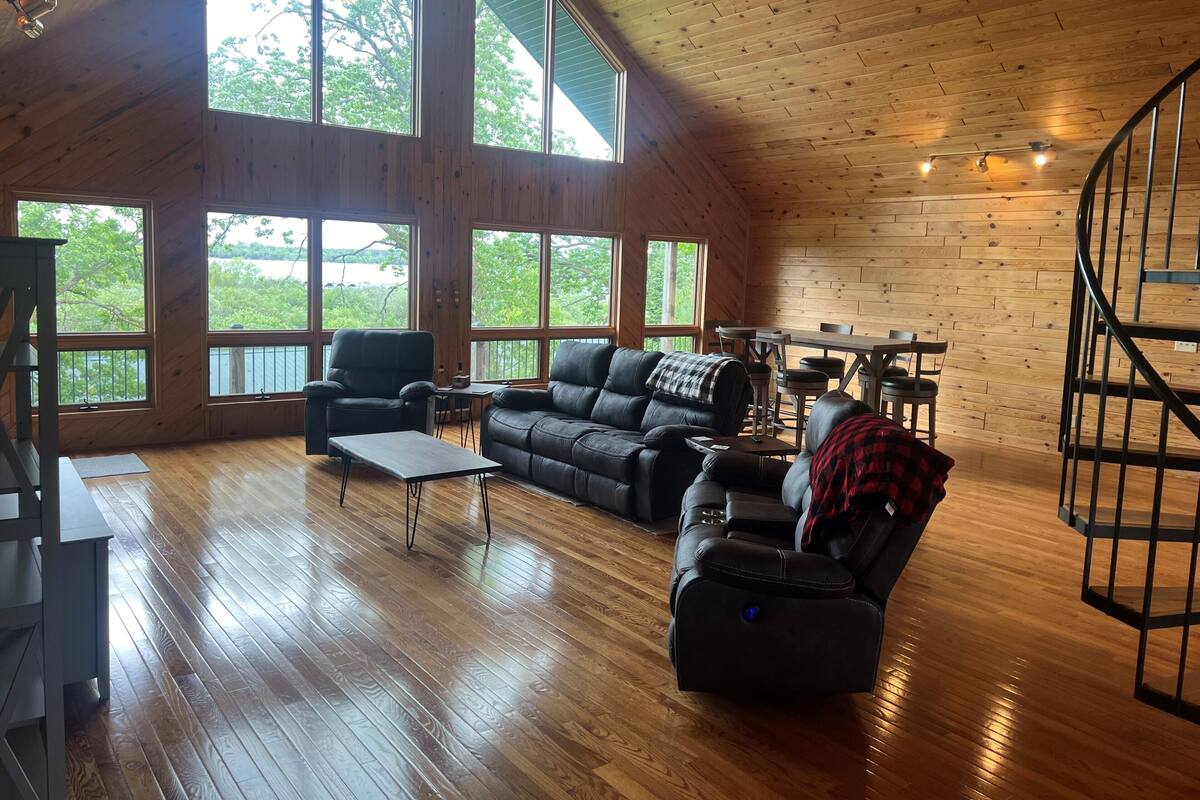 A spacious living room with wooden floors, large windows, a black leather sofa, a recliner, and a dining area with a table and chairs.
