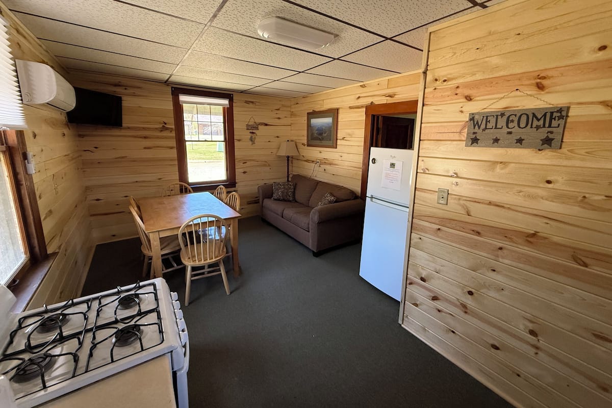 A cabin-style room with a wooden interior, a dining table with chairs, a sofa, a refrigerator, and a stove.