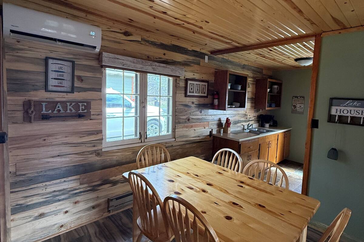 A wooden interior with a dining table and chairs, a kitchen area, and a sign that reads 'LAKE HOUSE.'