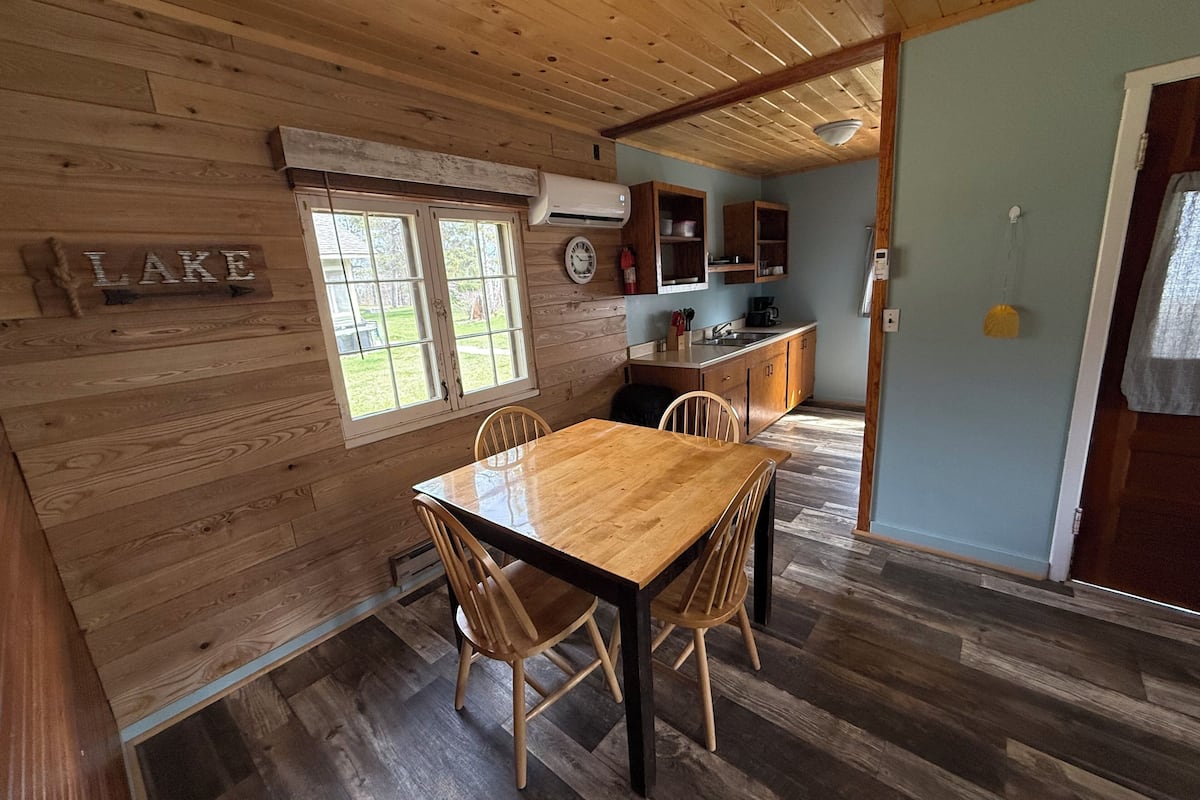 A wooden cabin interior with a dining table, chairs, a kitchen area, and a window with a view of the outdoors.